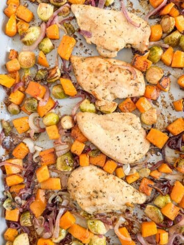 Roasted chicken breasts with sweet potatoes, Brussels sprouts, and caramelized onions on a baking sheet.